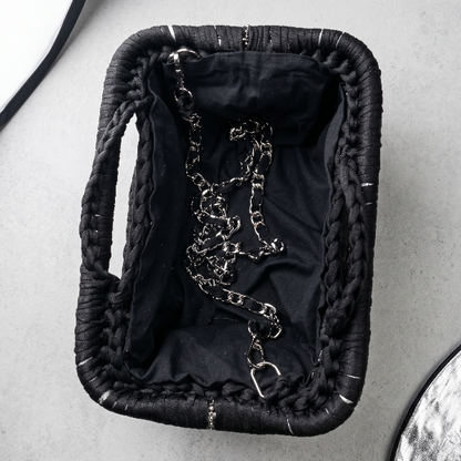A black woven basket with braided handles, shown from above on a light gray surface. The basket is lined with black fabric and contains a silver metal chain with a clasp.