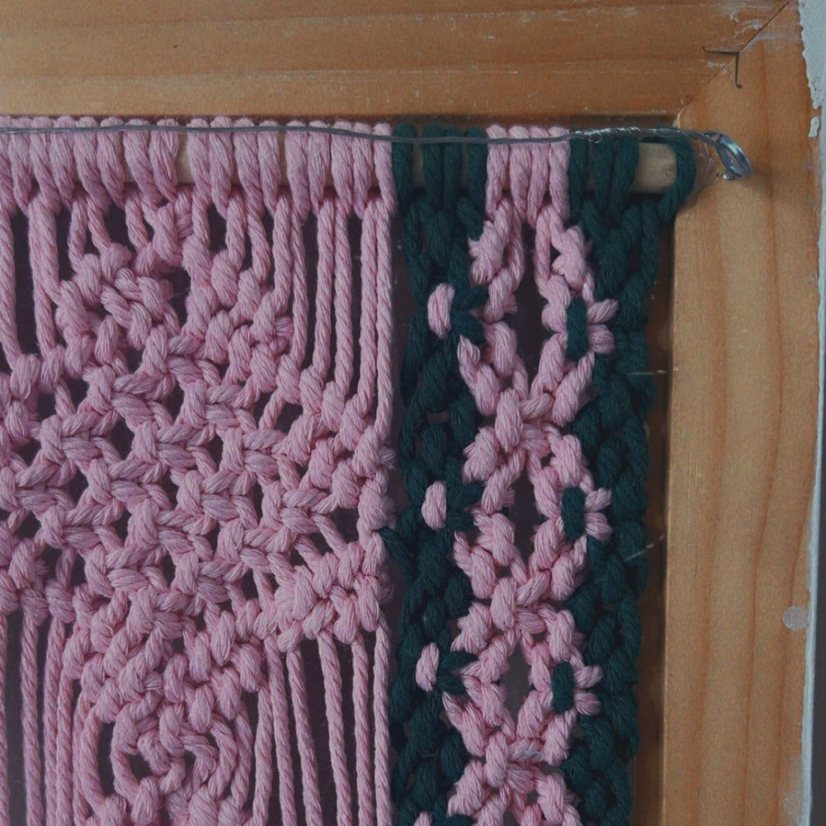 A close-up of a handmade woven wall hanging or textile panel with a pink openwork pattern and a dark green vertical border, mounted on a wooden frame.
